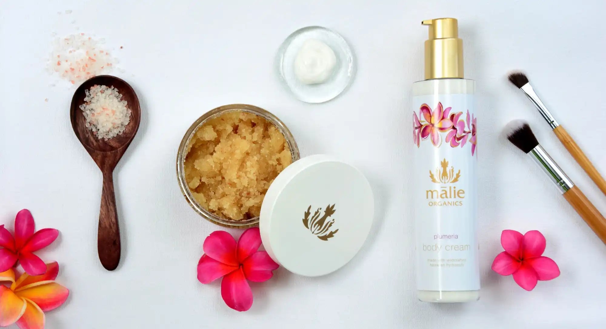 Beauty products and spa items arranged with pink plumeria flowers on a white surface.