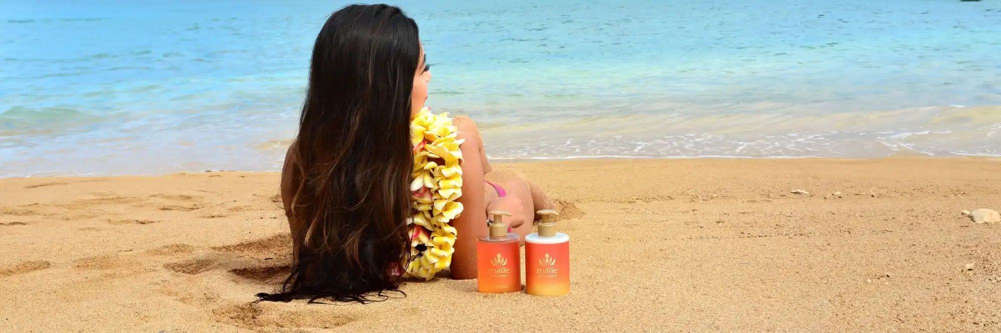 Someone with long dark hair wearing a yellow lei sits on a beach.