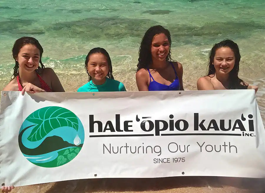 A white banner displaying ’Hale’opio Kaua’i’ with a green leaf logo and tagline ’Nurturing Our Youth Since 1975’’